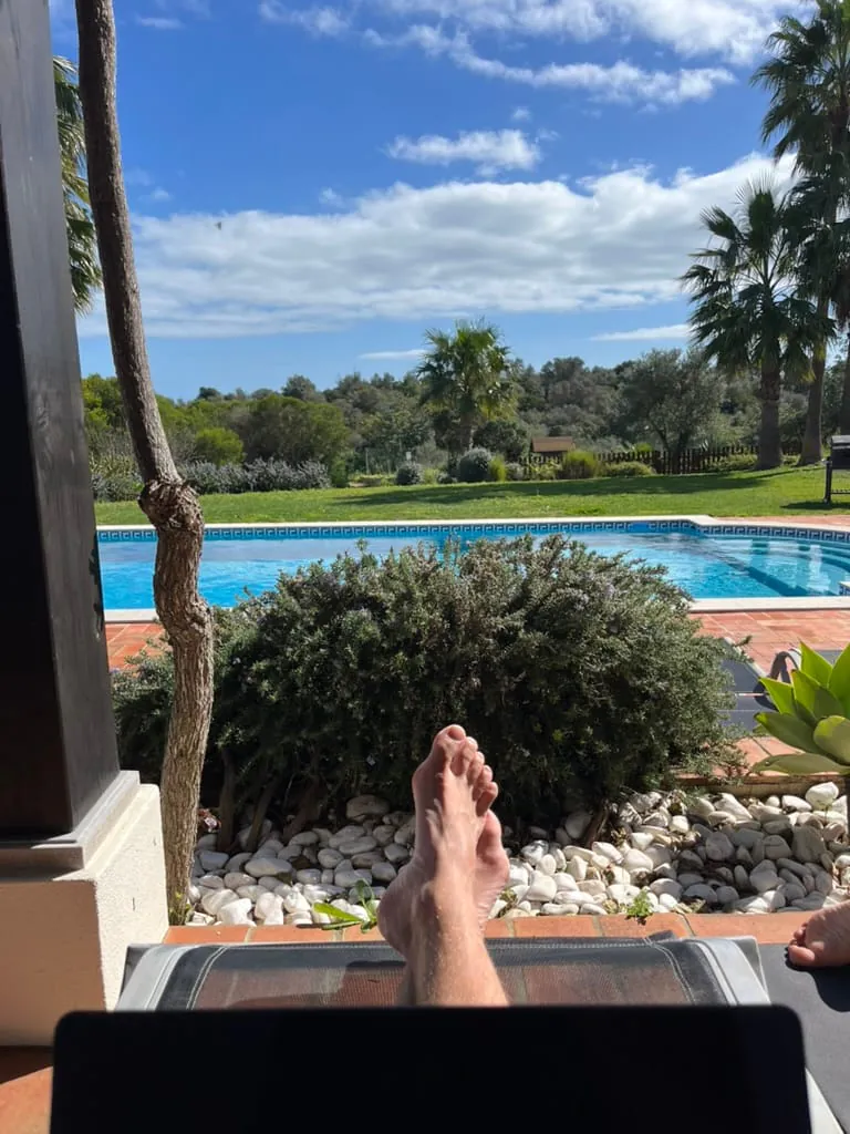 Bare feet crossed on a sun lounger seen over the top edge of an open laptop, the pool, lawn and palms in full sun beyond