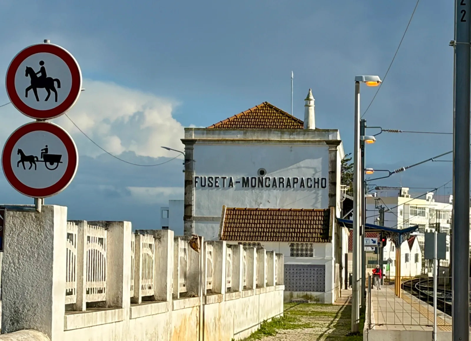 The whitewashed FUSETA-MONCARAPACHO station building seen along the platform, with overhead wires, street lamps and a single figure waiting by the track