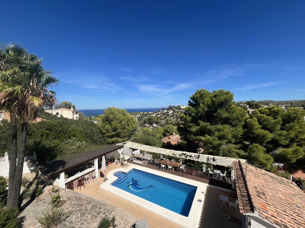 Looking down on a pool with a dolphin mosaic, an outdoor bar and a shaded terrace, pines and the sea beyond