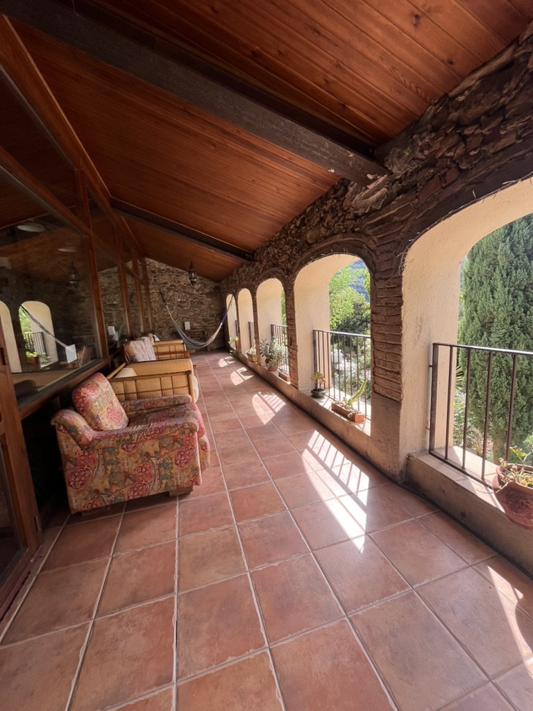 A tiled loggia under a wooden roof, stone arches open to the trees, armchairs and a hammock along the wall