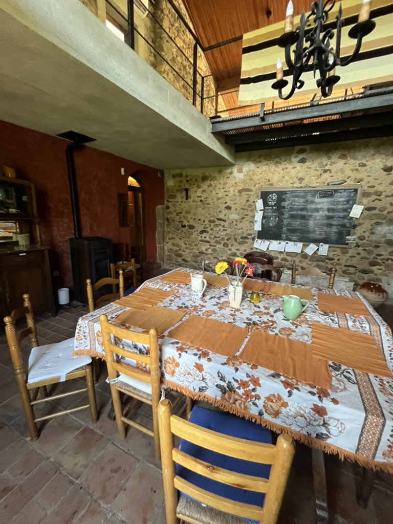 A long dining table with a floral cloth and flowers, wooden chairs, a stone wall with a chalkboard and a gallery overhead