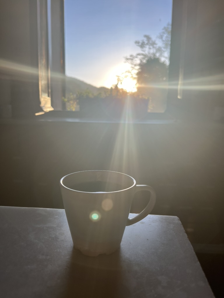 A white mug on a stone table in front of an open window, low sun breaking over wooded hills